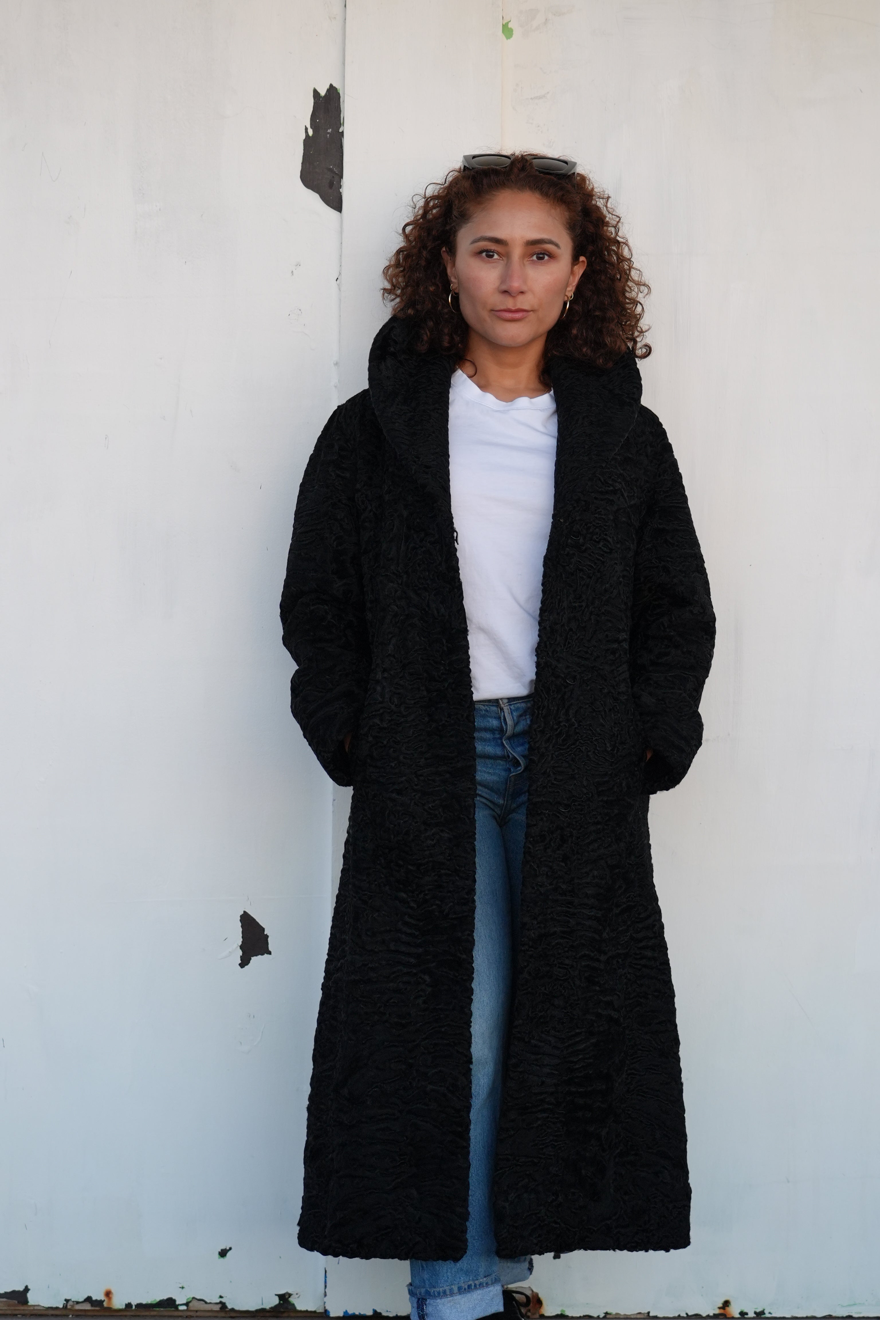 A woman with curly brown hair stands against a white wall wearing a long black Afghan karakul coat over a white t-shirt and blue jeans